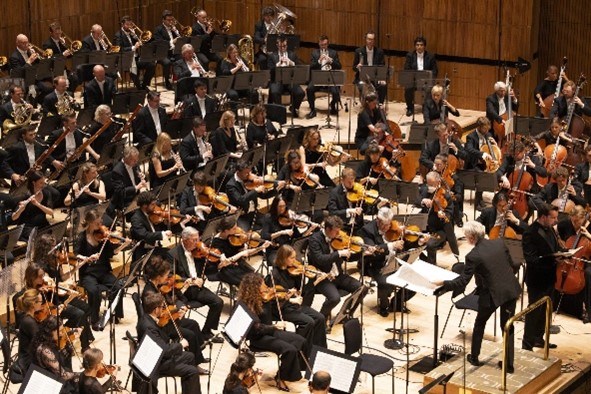 An orchestra is playing a piece of music, all facing the conductor, who commands the band in playing the piece.